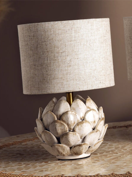Lámpara de sobremesa Piña beige de cerámica colocada sobre una mesa de mosaico, con base de hojas en relieve y pantalla de tejido natural.